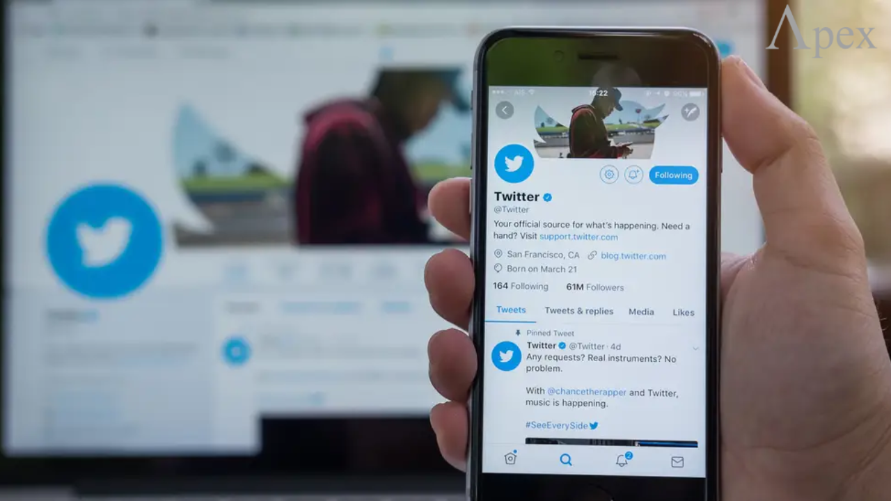 Person holding a smartphone displaying the official Twitter profile with its logo, followers count, and tweets, in front of a blurred laptop screen.